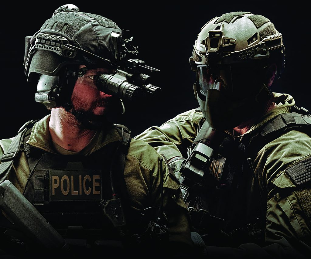 Two tactical officers in full gear with helmets, night vision goggles, face masks; one vest labeled 'POLICE'; both stand side by side, ready for action
