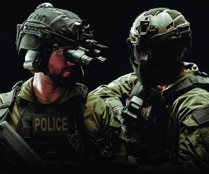 Two tactical officers in full gear with helmets, night vision goggles, face masks; one vest labeled 'POLICE'; both stand side by side, ready for action