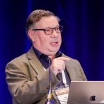 Person in brown suit stands at podium with IACP sign, holding microphone; open laptop on podium; blue curtain backdrop fills background