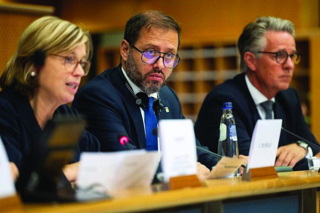 Three individuals in formal attire seated at conference table with microphones, nameplates, documents, and water bottle, suggesting official discussion