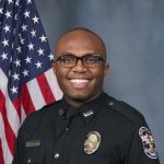 Police officer in formal dark uniform with badge and nameplate stands in front of an American flag backdrop, wearing a tie and visible shoulder insignia