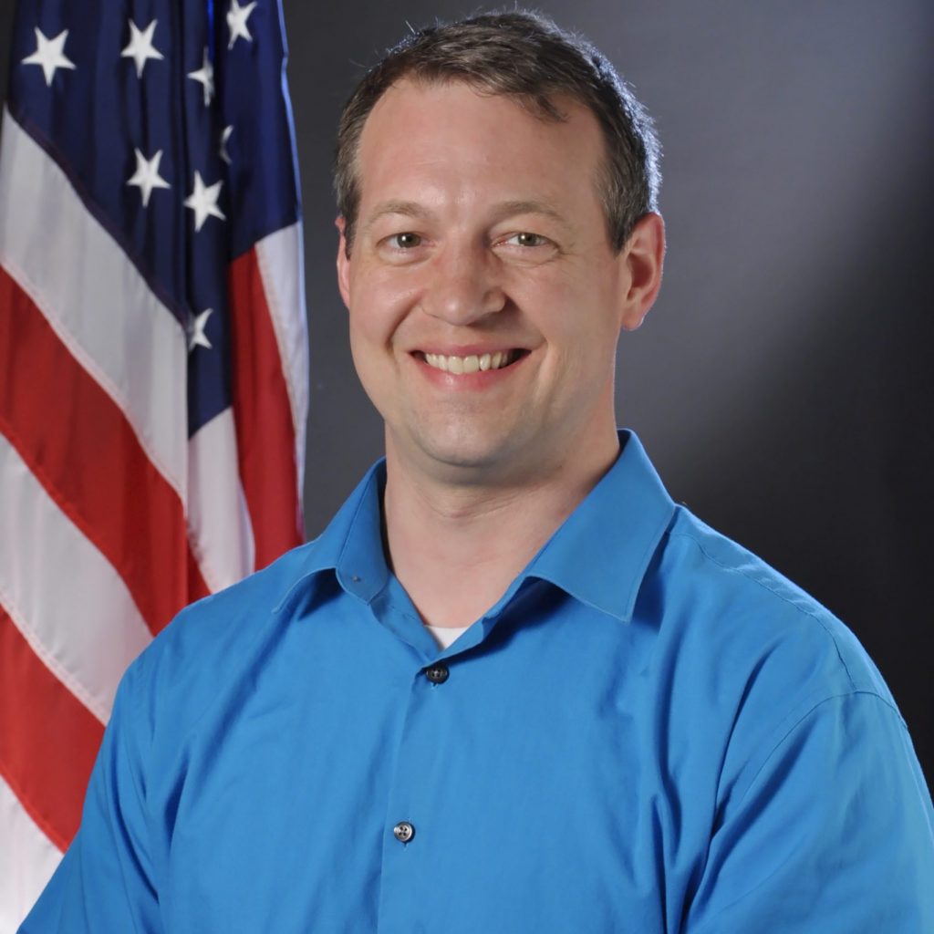 Person in bright blue button-up shirt sits before American flag; dark background emphasizes attire and setting