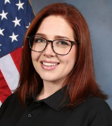 Person with shoulder-length red hair wearing a black collared shirt poses before a gray backdrop with a partially visible American flag to the left.