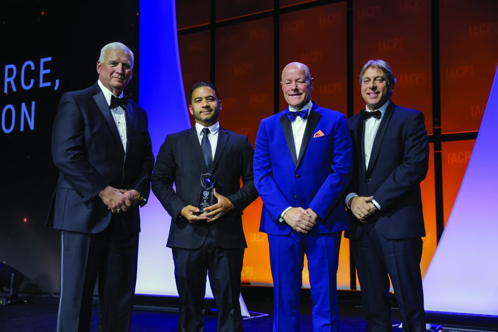 Five individuals in formal suits stand on stage with orange IACP backdrop; one person holds a glass award, others pose under bright event lighting.