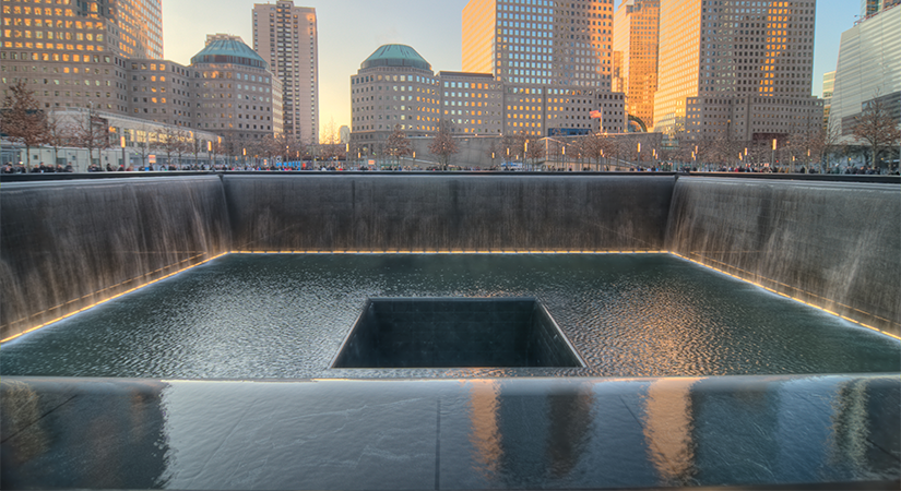 National September 11 Memorial reflecting pool features cascading waterfalls surrounding a central square void, bordered by engraved parapets, with surrounding trees, plaza walkways, and Lower Manhattan office towers illuminated by late-afternoon sunlight in the background