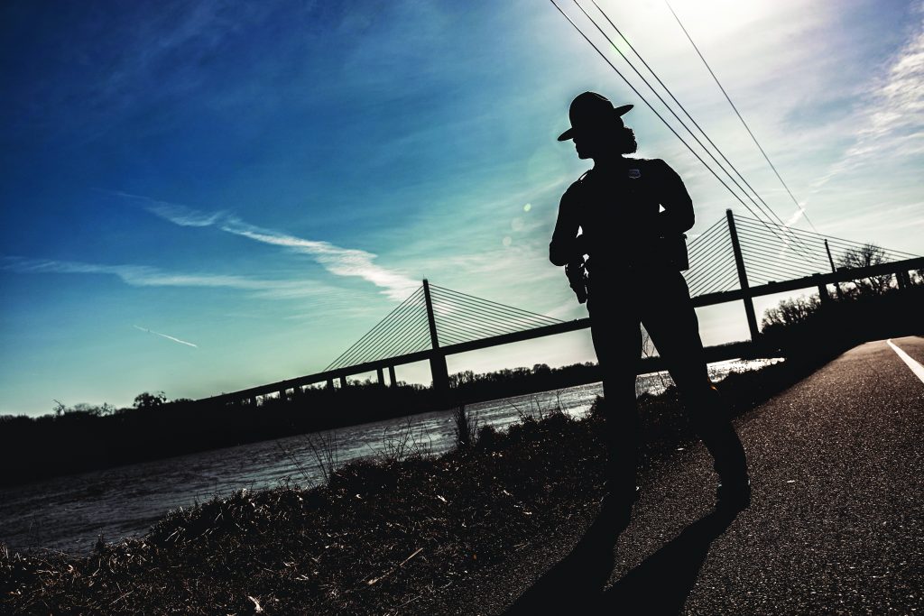 The image shows a silhouette of a police officer standing on the side of a road with hands on hips. The officer is wearing a wide-brimmed hat and uniform, with visible equipment on the belt. In the background, there is a large suspension bridge spanning across a body of water. The sky is clear with some wispy clouds, and power lines run parallel to the road. The scene is set during either sunrise or sunset, casting long shadows and creating a dramatic contrast between the dark figure and the bright sky. The officer's face is not visible due to the silhouette effect.