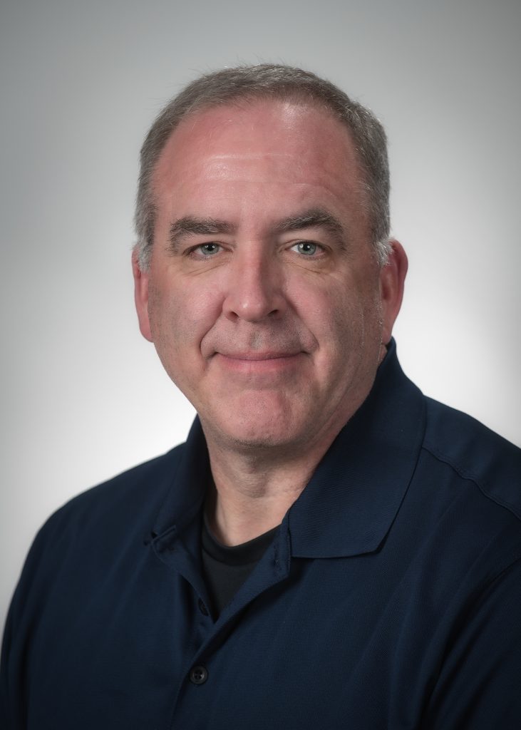 Portrait‑style image shows an individual wearing a dark collared shirt against a neutral gradient background, with shoulders and upper torso visible