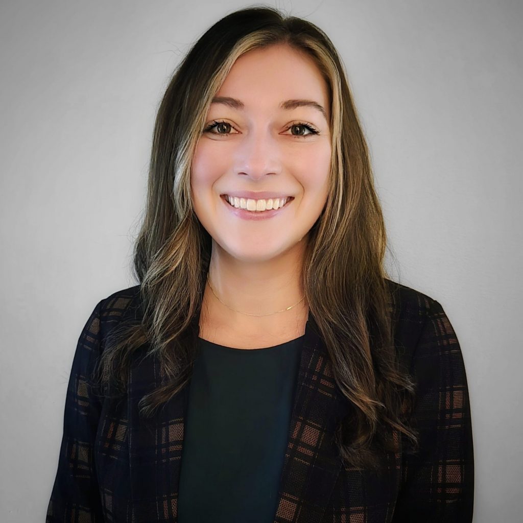 Individual with long wavy hair in dark plaid blazer over black top stands against plain gray background; face blurred to maintain anonymity.