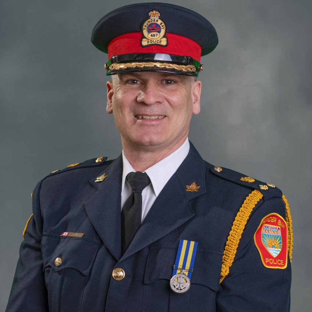 Officer in dark blue dress uniform with gold buttons, medals, 'Police' patch, and peaked cap with red-green bands—formal attire for official duty