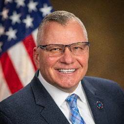 Individual in dark suit, white shirt, blue plaid tie stands before blurred American flag with brown backdrop