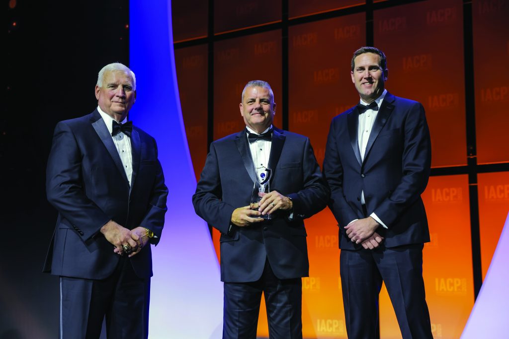 Three individuals in black tuxedos stand on stage with orange IACP backdrop; center person holds a glass award while others pose under bright event lighting.