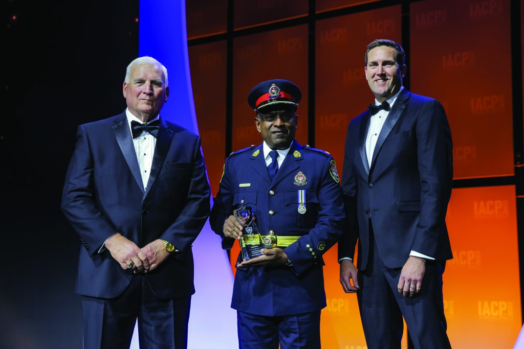 Three individuals in formal attire stand on stage with orange IACP backdrop; center person in decorated dress uniform holds a glass award under bright lights.