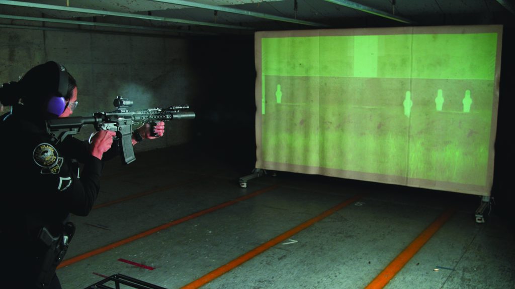A person in a police uniform is aiming a rifle at a large screen in an indoor shooting range. The screen displays green silhouettes of human figures for target practice. The person is wearing ear protection and appears to be in the process of firing the weapon, as there is visible smoke near the muzzle of the rifle. The environment is dimly lit with concrete walls and floor markings indicating lanes