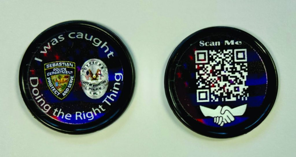 The image shows two round tokens placed on a white surface. The token on the left has a black background with three logos: the first logo is of the "Nassau County Police Department," the second logo is of an eagle with a shield, and the third logo is of another police badge. Surrounding these logos, there is text that reads, "I was caught Doing the Right Thing." The token on the right has a QR code in the center with text above it that says, "Scan Me!" Below the QR code, there are two hands shaking each other.