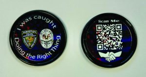 The image shows two round tokens placed on a white surface. The token on the left has a black background with three logos: the first logo is of the "Nassau County Police Department," the second logo is of an eagle with a shield, and the third logo is of another police badge. Surrounding these logos, there is text that reads, "I was caught Doing the Right Thing." The token on the right has a QR code in the center with text above it that says, "Scan Me!" Below the QR code, there are two hands shaking each other.