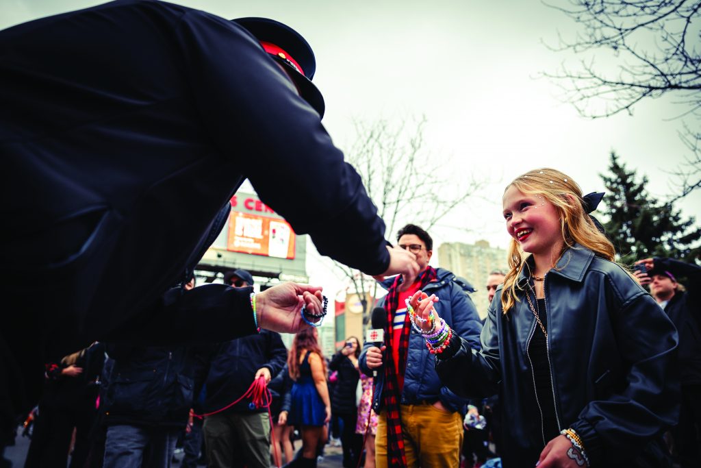 Person in black jacket and hat hands item to another in dark jacket; crowd gathered outdoors with trees and buildings in background