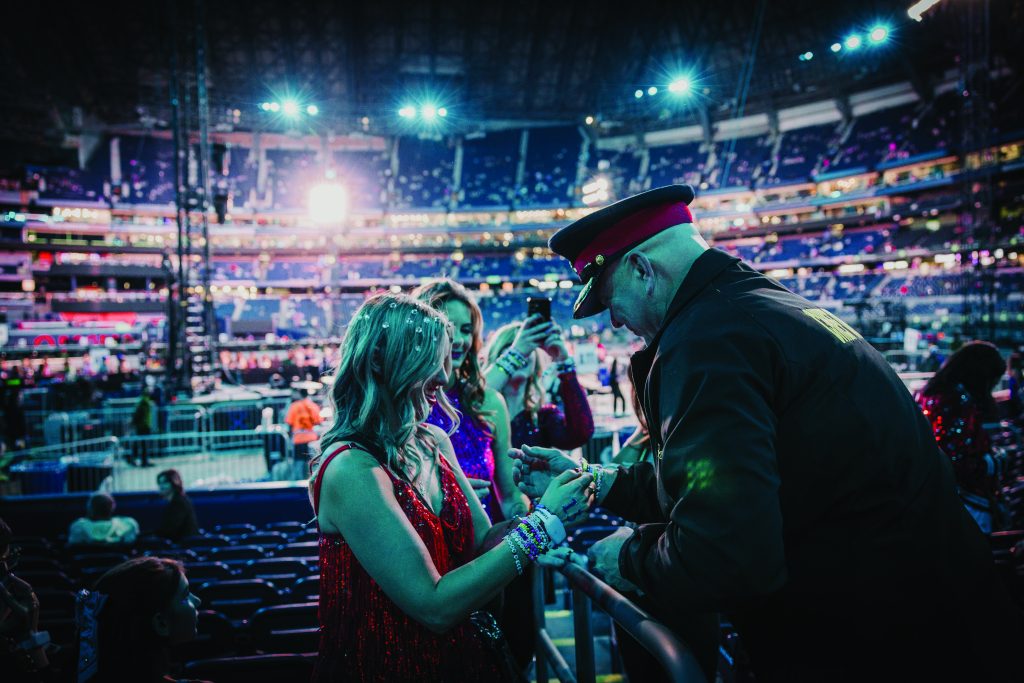 Police officer in uniform checks wristbands of three women in red and purple dresses at a crowded, brightly lit indoor venue with tiered seating