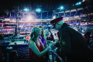 Police officer in uniform checks wristbands of three women in red and purple dresses at a crowded, brightly lit indoor venue with tiered seating