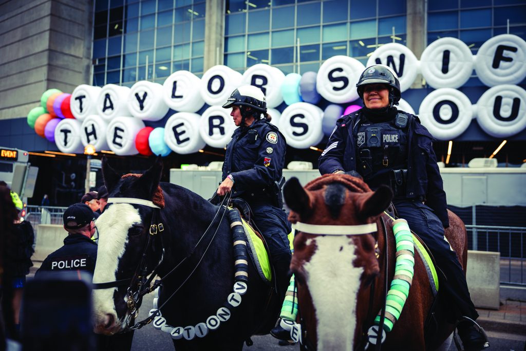 Mounted police patrol outside venue with large white balloons spelling “TAYLOR SWIFT THE ERAS TOUR”; horses wear decorative reins; crowd not visible