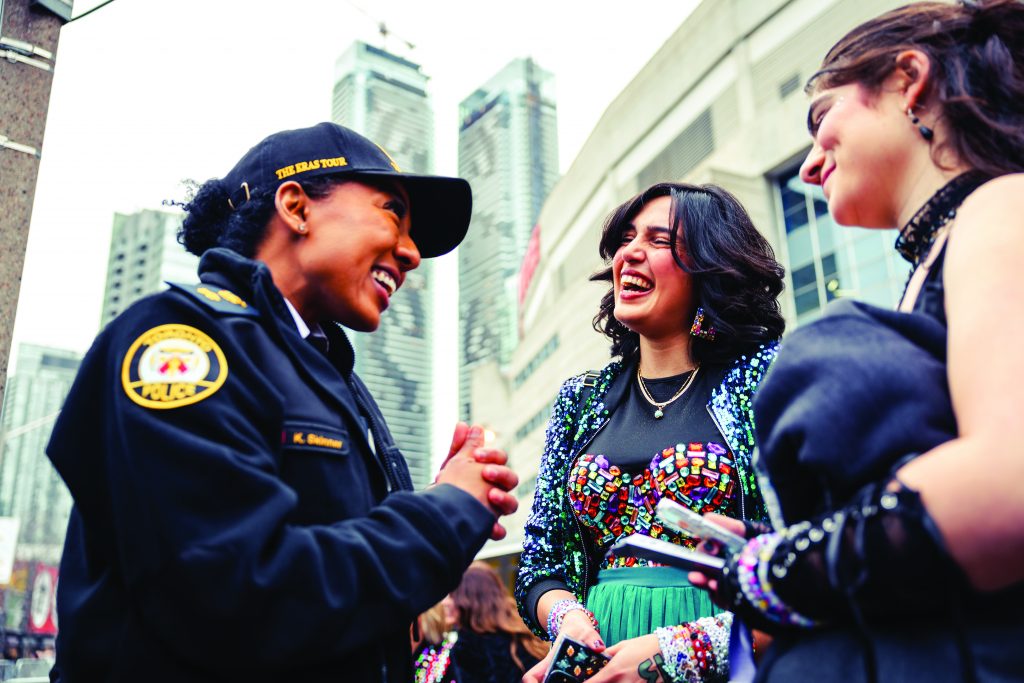 Uniformed officer speaks with two women in colorful concert attire; background features tall buildings and modern architectural structure