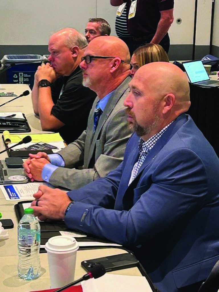 Participants sit at a conference table with microphones, papers, laptops, water bottles, and coffee cups, listening during a formal meeting session