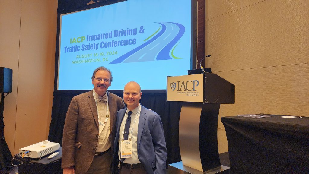 Two people stand near screen showing 'IACP Impaired Driving & Traffic Safety Conference, Aug 16–18, 2024, Washington, DC'; IACP podium at right