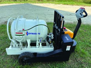 Dynasafe unit with white cylindrical tank on black wheeled base; gauges, hoses, control panel visible; placed on grass near gravel path.