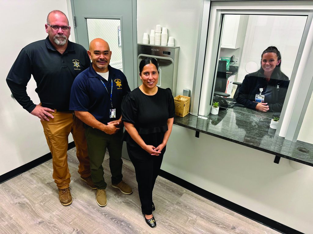 Four individuals stand in room with light wood flooring, granite service counter, door with window, and shelf holding stacked cups and assorted items
