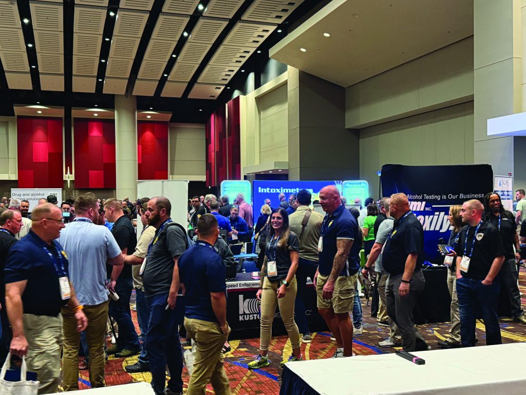 Convention hall with law enforcement professionals engaging at booths like Kustom Signals and Intoximeters, beneath high industrial ceiling