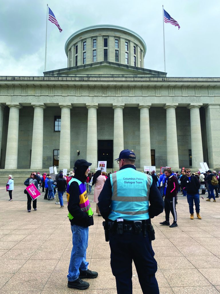 Crowd gathers with signs on a plaza in front of a stone capitol building with columns and dome, as an officer in a blue dialogue vest engages nearby participants