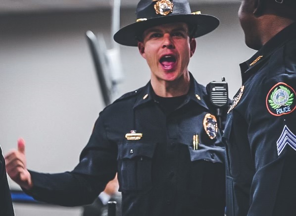 Two uniformed police officers in dark dress uniforms with badges, shoulder patches, and rank insignia, one wearing a campaign hat and radio on shoulder