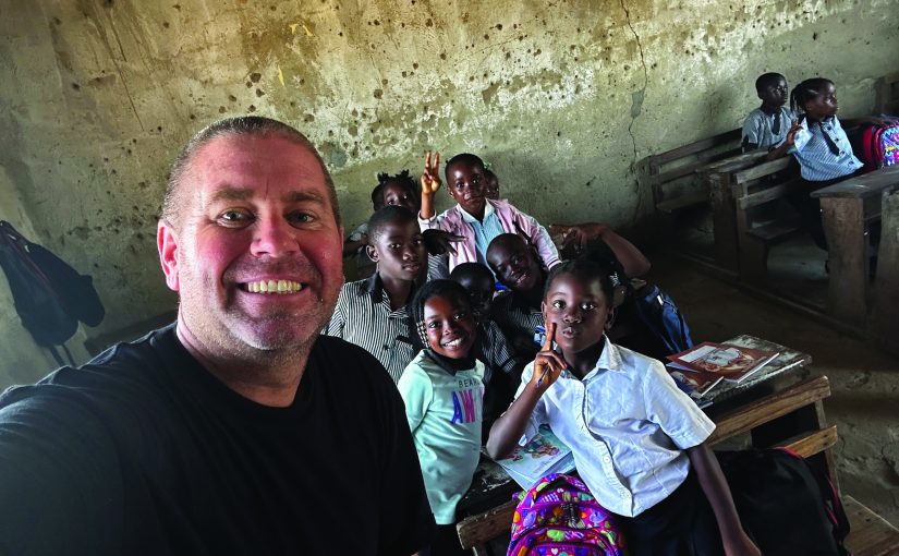 Visitor takes a group selfie with schoolchildren inside a worn classroom with wooden desks, backpacks, books, cracked walls and bare concrete floors