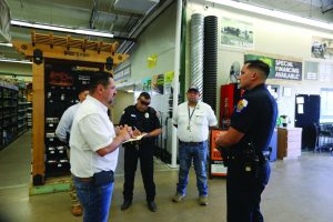 Group of men standing inside a hardware store near shelves and signage, including a police officer in uniform talking with others holding papers