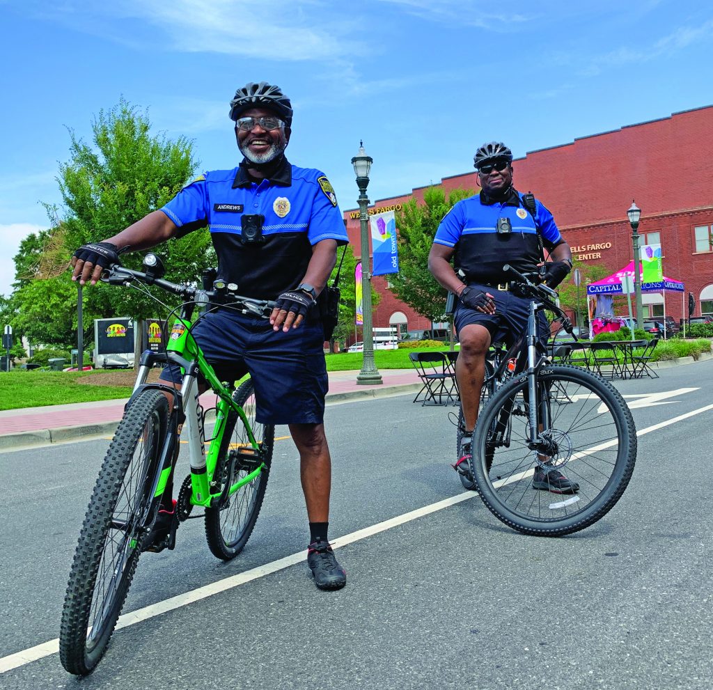 Two uniformed police officers wearing helmets and body cameras patrol on bicycles along a city street with brick building, banners, and outdoor tables in view