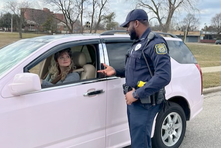 Police officer in uniform stands beside light pink SUV, speaking to driver through open window on suburban street with leafless trees and buildings behind