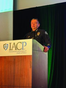 A police officer is standing behind a wooden podium with the IACP (International Association of Chiefs of Police) logo on it. The officer is wearing a dark uniform with various insignias and badges, including a shoulder patch and name tag. The officer's face is obscured by a solid color block for privacy. The background consists of dark green curtains and part of a white wall or screen.