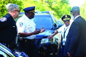 Four uniformed officers and a suited man stand near two police vehicles, discussing operations; trees and a second cruiser are visible in the background