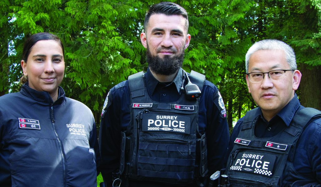 Three Surrey Police officers wearing black tactical uniforms and vests with visible name tags and Canadian flag patches, standing outdoors in front of green trees