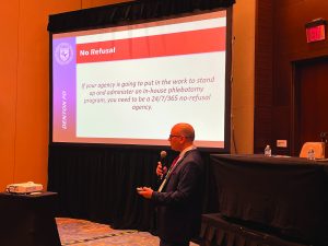 Presenter in suit stands before 'No Refusal' slide on in-house phlebotomy; beige room with table holding water bottles and law enforcement gear