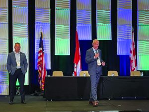 Two suited men speak on stage at a conference; behind them are U.S., Canadian, and another flag, with a backdrop of blue-lit vertical panels.