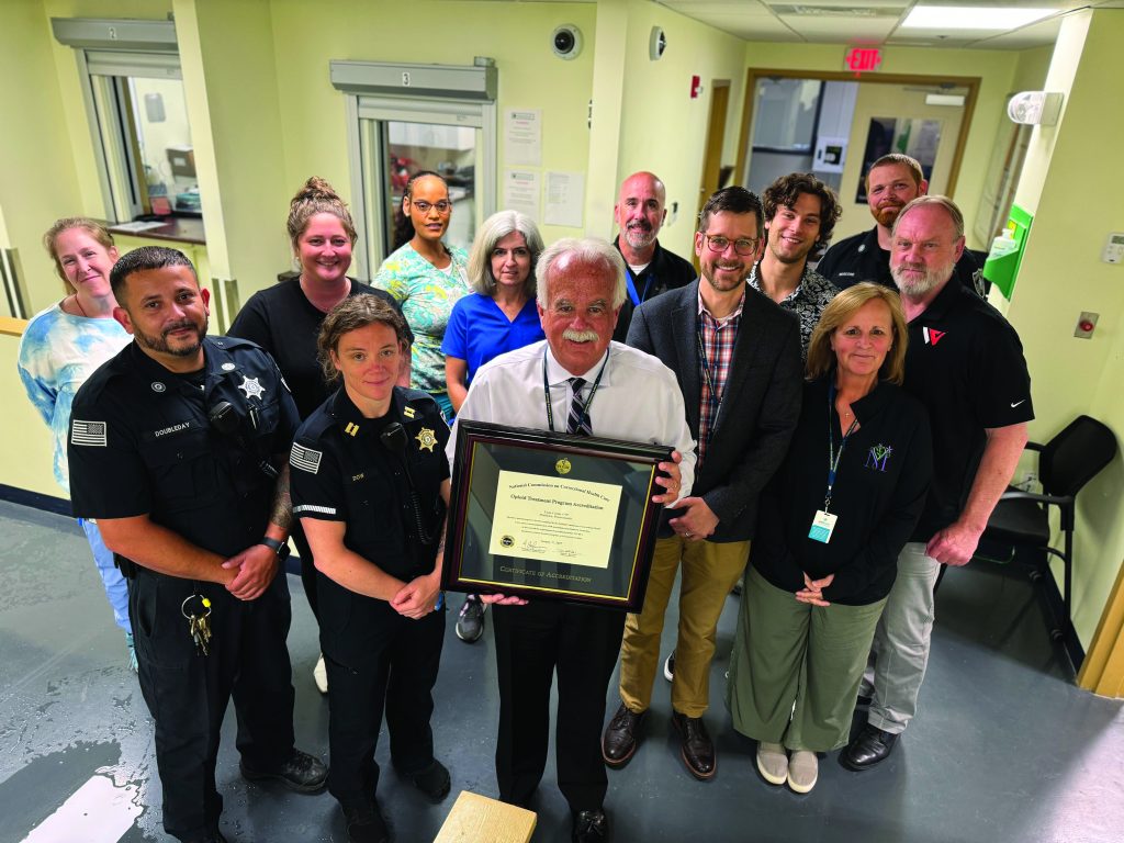 Group of law enforcement professionals and staff in hallway; one holds framed certificate. Background shows doors, windows, and office equipment