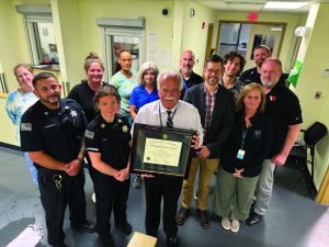Group of law enforcement professionals and staff in hallway; one holds framed certificate. Background shows doors, windows, and office equipment