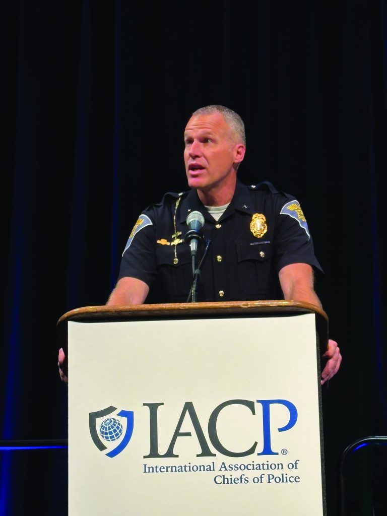 Uniformed police officer speaks at podium with IACP logo; dark background emphasizes formal setting, highlighting leadership in law enforcement