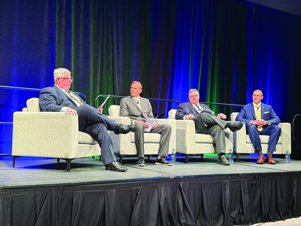 Four men in suits seated on stage in gray armchairs, holding papers and water bottles; dark curtain backdrop lit with green and blue stage lighting