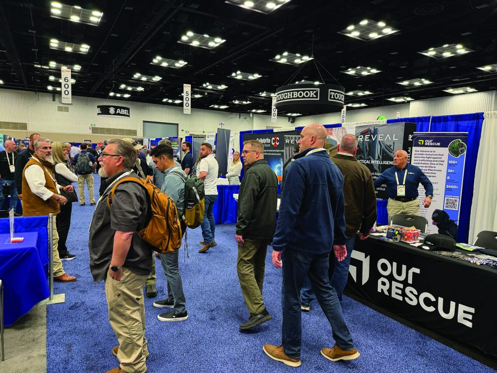 Busy convention hall with law enforcement professionals visiting booths like 'OUR RESCUE' and 'TOUGHBOOK'; blue carpet, bright overhead lighting