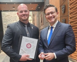 Two men in suits stand indoors; one holds a binder labeled 'Military Veterans Wellness Program,' the other displays a coin with the same program logo