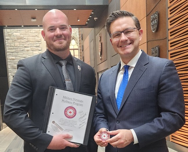 Two men in suits stand indoors; one holds a binder labeled 'Military Veterans Wellness Program,' the other displays a coin with the same program logo