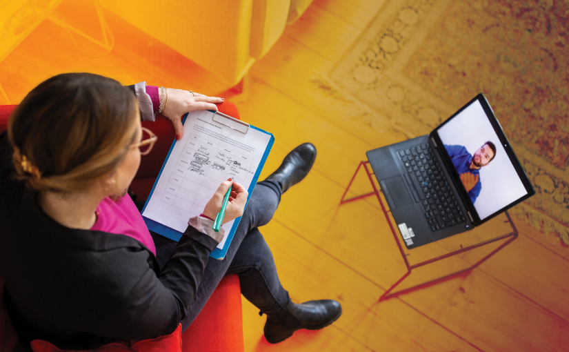 Person seated on red chair with clipboard, engaged in video call on laptop atop small table; room features wooden floor and patterned rug