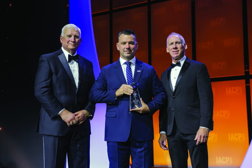 Three individuals in formal suits stand on stage with orange IACP backdrop; center person holds a glass award while others pose under bright event lighting.