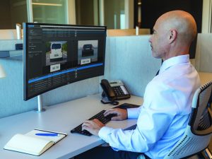 Person in blue shirt at desk with curved monitor showing vehicle images and license plates; using keyboard and mouse; phone and notebook nearby